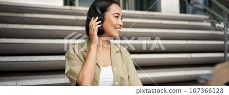 Close up portrait of smiling asian woman listens music in headphones, turns around with happy face expression, resting outdoors 107366128