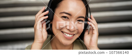 Close up portrait of smiling asian girl in headphones, listens to music outdoors, looking happy. 107366140