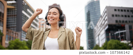 Happy asian woman in headphones, listening music and dancing on street of city centre, smiling with hands up 107366142