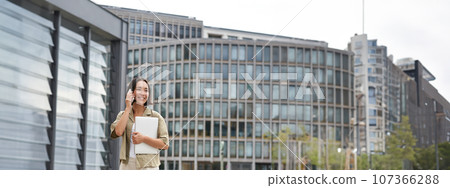 Stylish urban girl walking on street, talking on mobile phone and carrying laptop. Young woman making a telephone call while going somewhere Stylish urban girl walking on street, talking on mobile phone and carrying laptop. Young woman making a telephone call while going somewhere 107366288