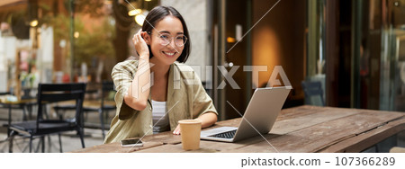 Portrait of smiling girl in glasses, sitting with laptop in outdoor cafe, drinking coffee and working remotely, studying online Portrait of smiling girl in glasses, sitting with laptop in outdoor cafe, drinking coffee and working remotely, studying online 107366289