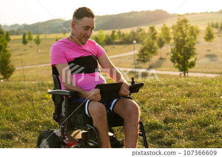 Gay man in wheelchair holds tablet on lap talking to boyfriend on videocall. Male with tattoo on neck enjoys talking on videocall in sunlit meadow 107366609