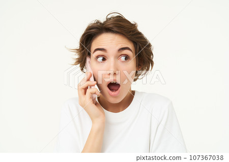 Portrait of amazed brunette girl looks surprised while talks on mobile phone, answers a call, amazed by big news, gasping shocked, standing over white background Portrait of amazed brunette girl looks surprised while talks on mobile phone, answers a call, amazed by big news, gasping shocked, standing over white background 107367038