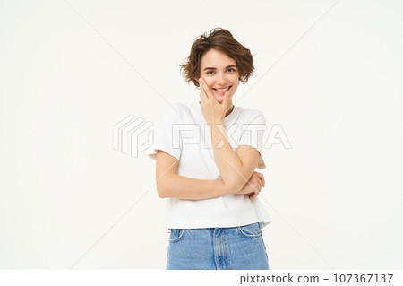 Skincare and women beauty concept. Portrait of young smiling woman, touches her face, gazing at camera, enjoys smooth facial skin after skincare routine, white studio background Skincare and women beauty concept. Portrait of young smiling woman, touches her face, gazing at camera, enjoys smooth facial skin after skincare routine, white studio background 107367137