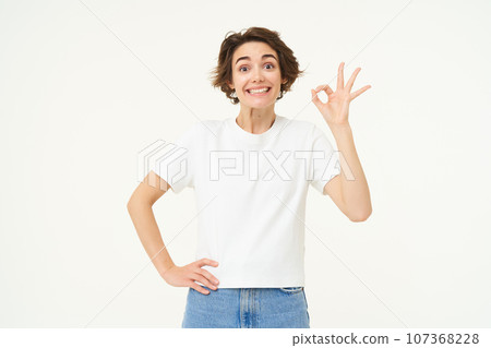 Portrait of woman smiling, showing okay sign with confidence, gives approval, recommends smth good, stands over white background 107368228