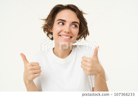 Close up shot of cheerful smiling girl, shows thumbs up, gives approval, likes something, positive feedback concept, stands over white background 107368233