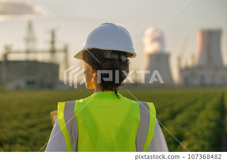 Back view of female engineer constructor stands on field on industrial enterprise background Back view of female engineer constructor stands on field on industrial enterprise background 107368482