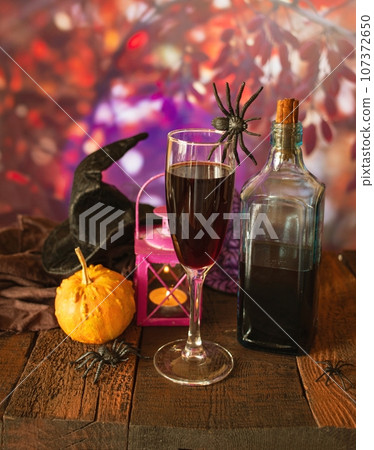 Halloween, red wine, lantern with a burning candle, pumpkins, spiders and a witch hat on a wooden table against the background of autumn nature, Halloween celebration Halloween, red wine, lantern with a burning candle, pumpkins, spiders and a witch hat on a wooden table against the background of autumn nature, Halloween celebration 107372650