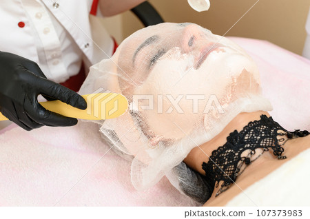 The patient's face is covered with gauze, the beautician applies cream to the girl's face with a spatula. The patient's face is covered with gauze, the beautician applies cream to the girl's face with a spatula. 107373983