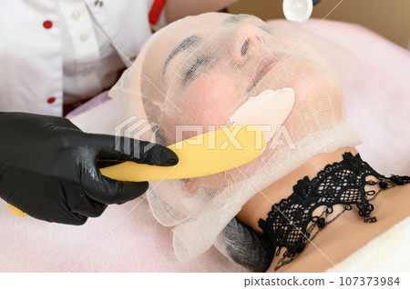 The beautician uses a spatula to apply the cream to the already prepared face with gauze. 107373984