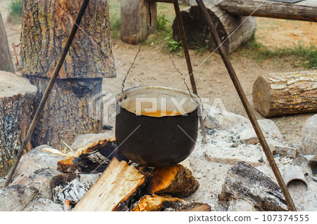 Metal cauldron with meal inside hanging on tripod over firewood, preparing for boiling on bonfire in village site. Metal cauldron with meal inside hanging on tripod over firewood, preparing for boiling on bonfire in village site. 107374555