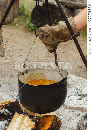 Hand of man in glove hanging cauldron with soup on hook of tripod over firewood, preparing for cooking on campfire. Hand of man in glove hanging cauldron with soup on hook of tripod over firewood, preparing for cooking on campfire. 107374556