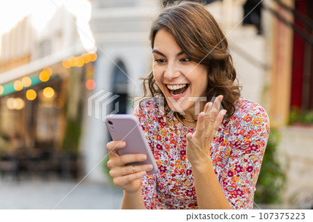 Happy woman use mobile smartphone celebrating win good message news outdoors in urban city street 107375223