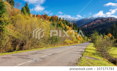straight road through mountainous countryside in autumn. trees on the hill in colorful foliage. grassy meadow on the roadside. warm sunny weather. old cracked asphalt 107375783
