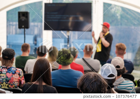 Male speaker with badge around neck talking into mic, while standing on stage and explaining business strategy to participants sitting in hall during presentation 107376477