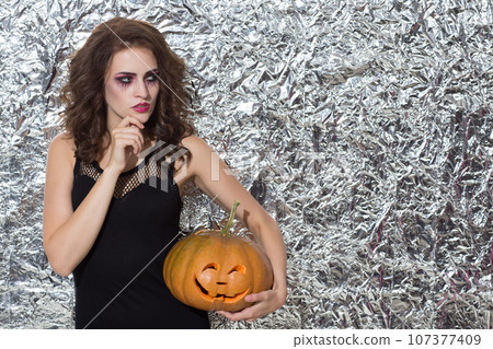 Pensive beautiful woman with decorative makeup, posing with pumpkin at Halloween. Front view of thoughtful female witch looking away, while plotting something, against foil wall. Concept of Halloween. Pensive beautiful woman with decorative makeup, posing with pumpkin at Halloween. Front view of thoughtful female witch looking away, while plotting something, against foil wall. Concept of Halloween. 107377409
