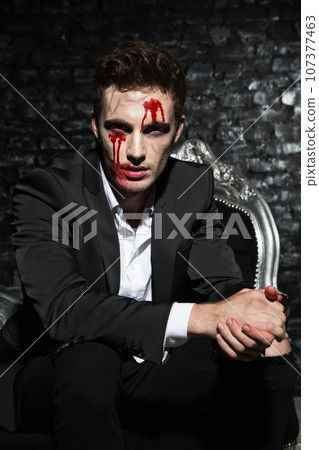 Portrait of a handsome young man sitting on a couch wearing a suit, with bloody make up on his face, looking serious with his hands crossed Portrait of a handsome young man sitting on a couch wearing a suit, with bloody make up on his face, looking serious with his hands crossed 107377463