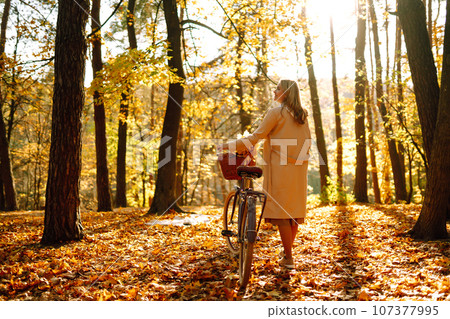 Stylish woman with a bicycle enjoying autumn weather in the park. Beautiful Woman in the autumn forest. 107377995