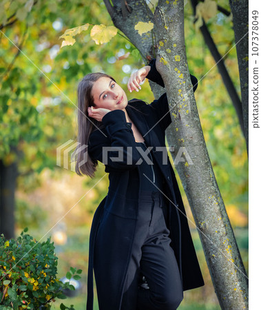 A beautiful girl stands by a tree in an autumn park. 107378049
