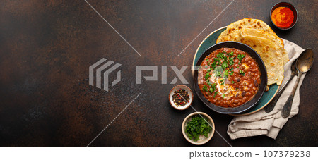 Traditional Indian Punjabi dish Dal makhani with lentils and beans in black bowl served with naan flat bread, fresh cilantro and two spoons on brown concrete rustic table top view. Space for text Traditional Indian Punjabi dish Dal makhani with lentils and beans in black bowl served with naan flat bread, fresh cilantro and two spoons on brown concrete rustic table top view. Space for text 107379238