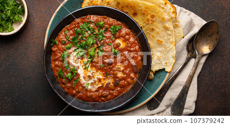 Traditional Indian Punjabi dish Dal makhani with lentils and beans in black bowl served with naan flat bread, fresh cilantro and two spoons on brown concrete rustic table top view 107379242