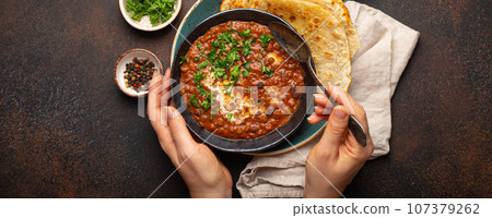 Female hands holding a bowl and eating traditional Indian Punjabi dish Dal makhani with lentils and beans served with naan flat bread, fresh cilantro on brown concrete rustic table top view 107379262
