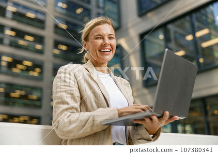 Smiling mature woman entrepreneur working laptop on modern building background and looks camera Smiling mature woman entrepreneur working laptop on modern building background and looks camera 107380341