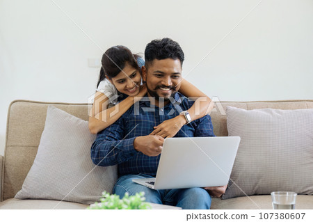 Happy loving family Indian couple hugging on couch at home and focused on laptop screen watching romantic movie Happy loving family Indian couple hugging on couch at home and focused on laptop screen watching romantic movie 107380607