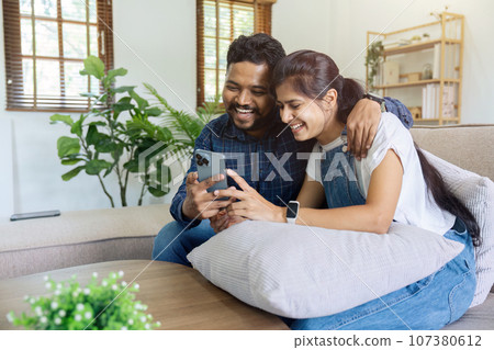 happy indian couple relax sitting on sofa at home holding phone video calling distance with family happy indian couple relax sitting on sofa at home holding phone video calling distance with family 107380612