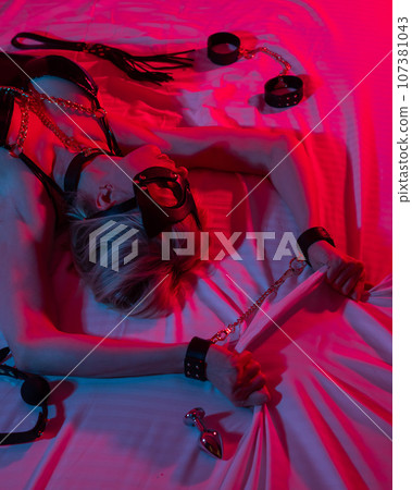 Sexy blonde woman in a leather mask and handcuffs and a collar lies in the bedroom in a red-blue neon light. Role-playing games. Sexy blonde woman in a leather mask and handcuffs and a collar lies in the bedroom in a red-blue neon light. Role-playing games. 107381043