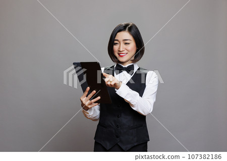 Smiling restaurant receptionist in uniform checking guest list on digital tablet. Eatery asian woman waitress tapping on touchscreen while examining online order while posing in studio Smiling restaurant receptionist in uniform checking guest list on digital tablet. Eatery asian woman waitress tapping on touchscreen while examining online order while posing in studio 107382186