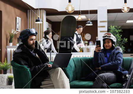 Boyfriend using laptop and girlfriend with mobile phone enjoying digital communication. Wintersports enthusiasts in hotel lobby for ski and snowboarding fun waiting with their smart devices. Boyfriend using laptop and girlfriend with mobile phone enjoying digital communication. Wintersports enthusiasts in hotel lobby for ski and snowboarding fun waiting with their smart devices. 107382283