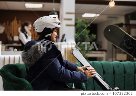 Asian female traveler in winter jacket comfortably seated on sofa with her ski equipment in hotel reception. Enthusiastic woman with snow helmet placing her skiing skis nearby in lounge area. 107382362