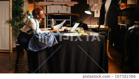 Seamstress using sewing machine to design and craft fashion items at workstation. Professional tailor working with handcraft equipment and tools, creating tailored custom made clothes. 107382366