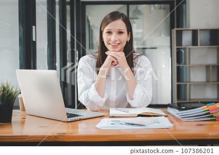 Portrait of young smiling woman looking at camera with crossed arms. Happy girl sitting in creative office. Successful businesswoman standing in office with copy space. 107386201
