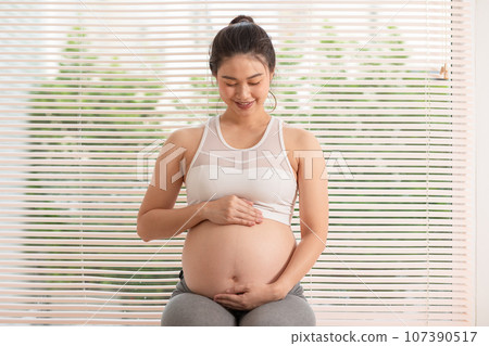Happy Pregnant Asian Woman sitting on a bed holding and stroking her big belly at home. Pregnancy of young woman enjoying with future life relax at home. Motherhood and Pregnant Concept. Soft focus Happy Pregnant Asian Woman sitting on a bed holding and stroking her big belly at home. Pregnancy of young woman enjoying with future life relax at home. Motherhood and Pregnant Concept. Soft focus 107390517