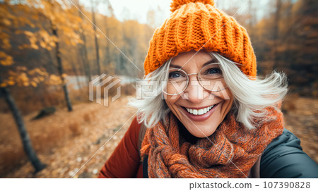 Happy older woman smiles takes selfie in autumn park. Positive cheerful woman in orange hat enjoying walk outdoors Happy older woman smiles takes selfie in autumn park. Positive cheerful woman in orange hat enjoying walk outdoors 107390828
