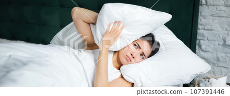 Insomnia. Asian girl shuts her ears with pillow as partner snoring. Woman lying in bed, bothered by noise at night, cannot sleep 107391446