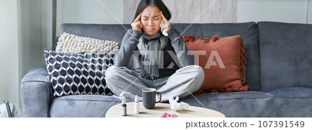 Young asian woman feels unwell, touches head, has headache and fever, staying at home on sick leave, drinking hot tea and taking drugs prescribed by GP Young asian woman feels unwell, touches head, has headache and fever, staying at home on sick leave, drinking hot tea and taking drugs prescribed by GP 107391549