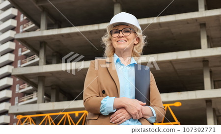 Positive businesswoman in helmet smiles against building 107392771