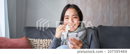 Portrait of sick woman contacting her GP via mobile phone while catching cold, sitting with flu at home, using smartphone 107395469