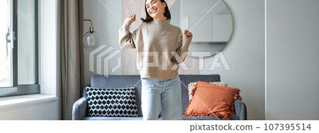 Enthusiastic asian woman dancing, feeling free and upbeat at her home, enjoying happy day, staying indoors, smiling and laughing 107395514