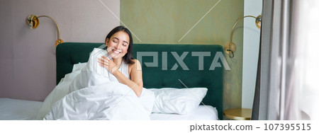 Happy korean woman wakes up in her hotel room, lying in bed under cozy warm blankets, white sheets, enjoying weekend morning 107395515