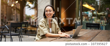 Portrait of asian girl works in cafe, uses laptop and sits outdoors on street. Digital nomad and online learning concept 107395627