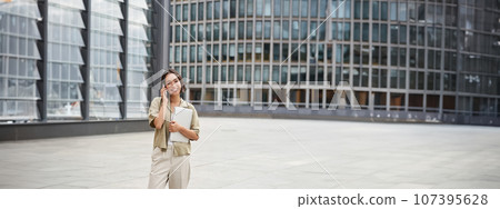 Stylish urban girl walking on street, talking on mobile phone and carrying laptop. Young woman making a telephone call while going somewhere 107395628