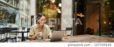 Young woman sitting on online meeting in outdoor cafe, talking to laptop camera, explaining something, drinking coffee 107395649