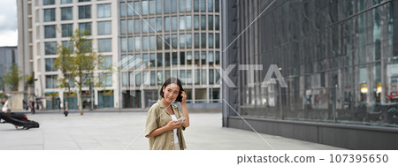 Portrait of asian woman walking in city. Street style shot of girl with smartphone, posing outdoors on street Portrait of asian woman walking in city. Street style shot of girl with smartphone, posing outdoors on street 107395650