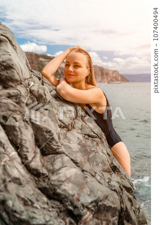 Woman swimsuit sea. Attractive blonde woman in a black swimsuit enjoying the sea air on the seashore around the rocks. Travel and vacation concept. 107400494