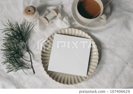 Christmas stationery. Blank square greeting card, invitation mockup, ceramic plate. Silk ribbon and pine tree branches. White linen tablecloth. Cup of coffee, winter breakfast. Festive holiday flatlay 107406629