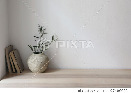 Moody Mediterranean home design. Vintage vase with olive tree branches. Old books on wooden table. Living room still life, home office. Empty white wall copy space. Modern interior, no people. 107406631